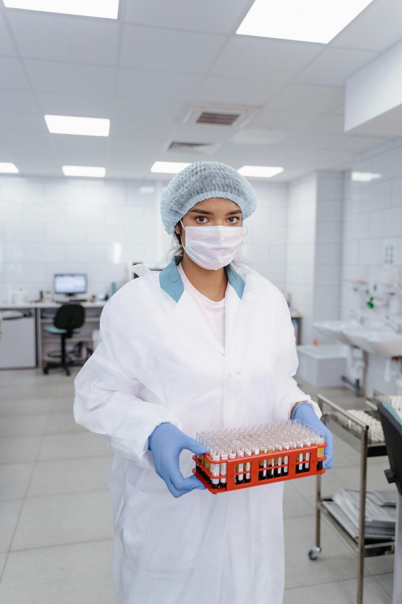 A scientist in protective gear holds a tray of test tubes in a modern laboratory setting.